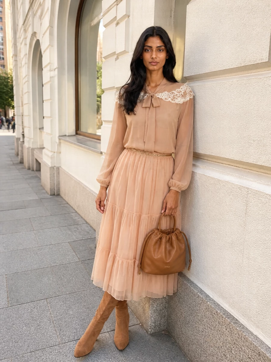 A gossamer tiered skirt and sheer lace-trim blouse in nude tones create a soft, romantic column of color that feels quietly luxurious for early autumn. Camel suede over-the-knee boots ground the lightness with warm texture, while a caramel bucket bag and fine gold chain belt introduce just enough structure and shine. The result is a fluid, boho-leaning ensemble that moves easily from brunch to softly polished dinners.