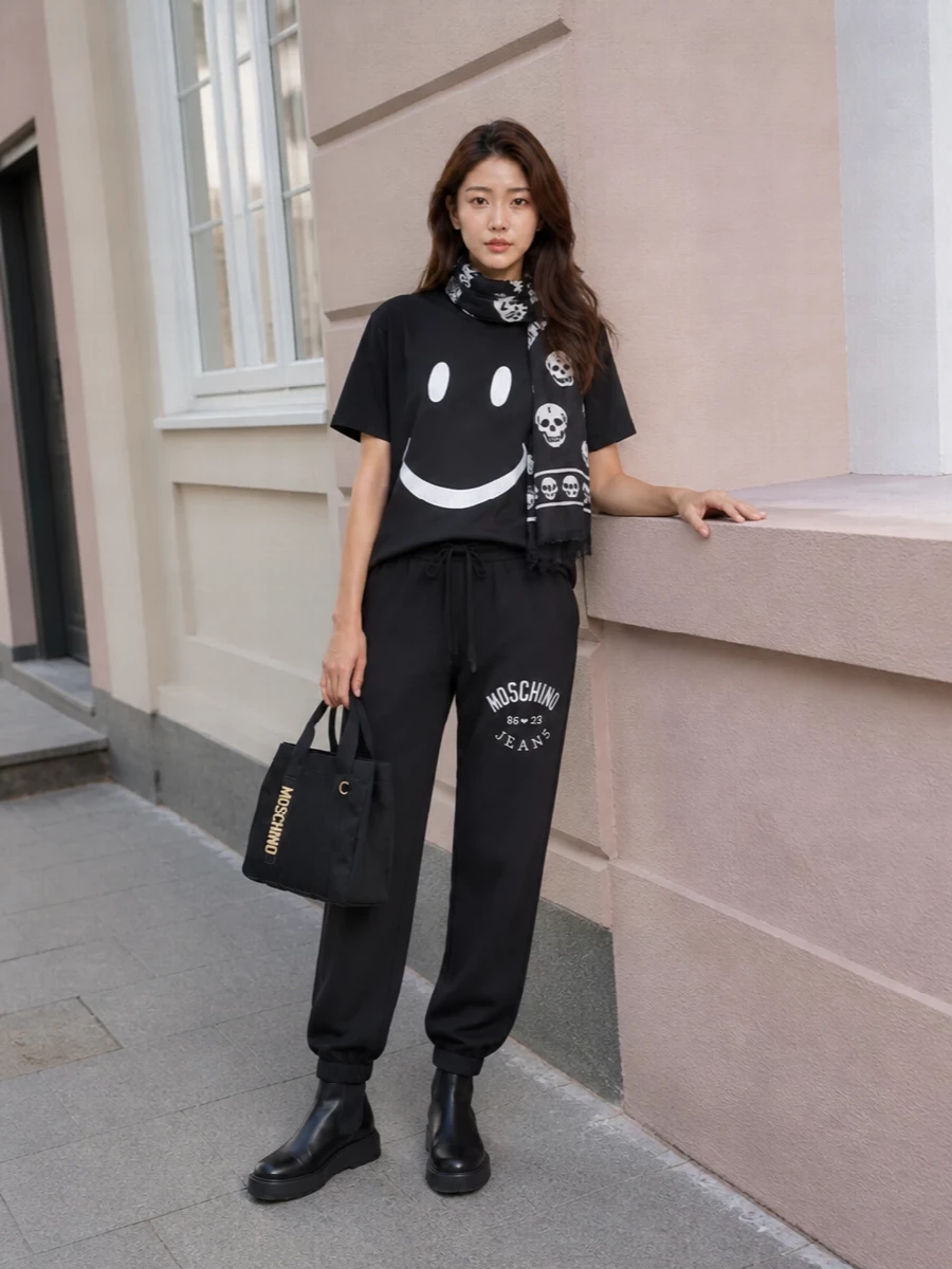 Head-to-toe black keeps the Moschino smiley tee sharp, pairing it with relaxed logo joggers for a graphic, off-duty street set. Chunky slip-on boots ground the proportions, while a compact black Moschino tote and monochrome skull scarf add branded edge without stealing focus from the tee. It is an easy city uniform for cooler days that still feels witty and fashion-aware.