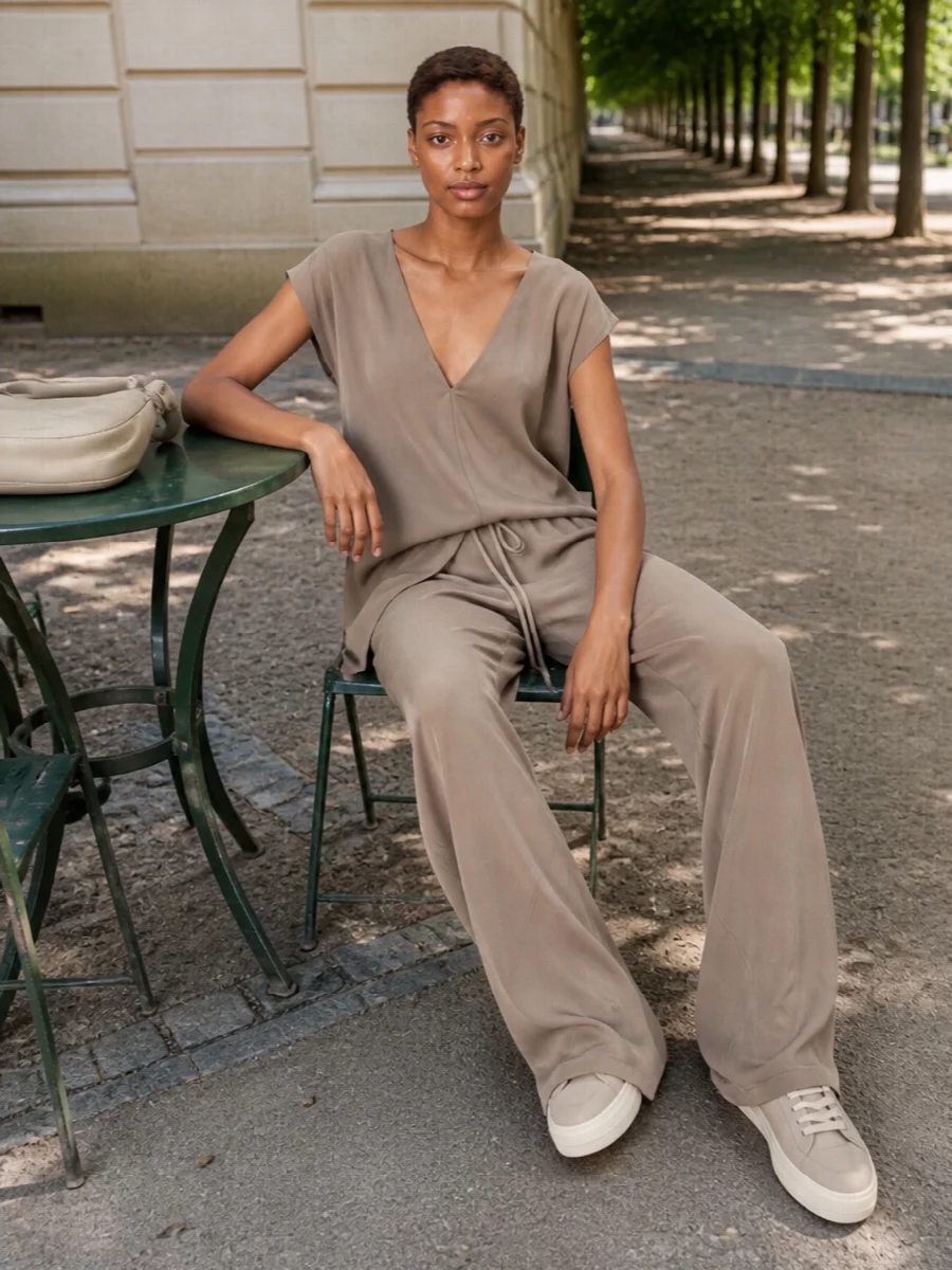 A taupe V-neck top falls into fluid high-waisted wide-leg trousers, reading as a single, elongated column in soft neutrals. Streamlined sneakers keep it grounded and relaxed, while the sculpted ivory shoulder bag adds just enough structure for city days or quiet dinners. The result is a calm, minimalist uniform with subtle Rick Owens attitude rather than overt drama.