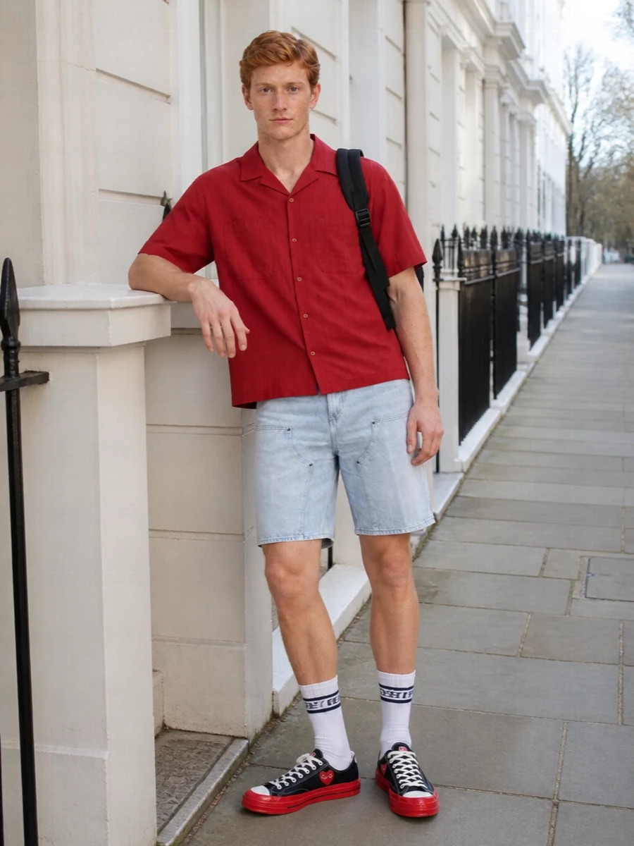 A brick-red camp shirt sits over light-wash carpenter shorts for a relaxed, skate-ready take on warm weather dressing. Graphic black-and-red canvas sneakers and collegiate striped socks pick up the color story without feeling forced, while a black utility backpack grounds everything in practical city wear. The result is easy, preppy-inflected streetwear that feels considered but not precious for day-long movement.