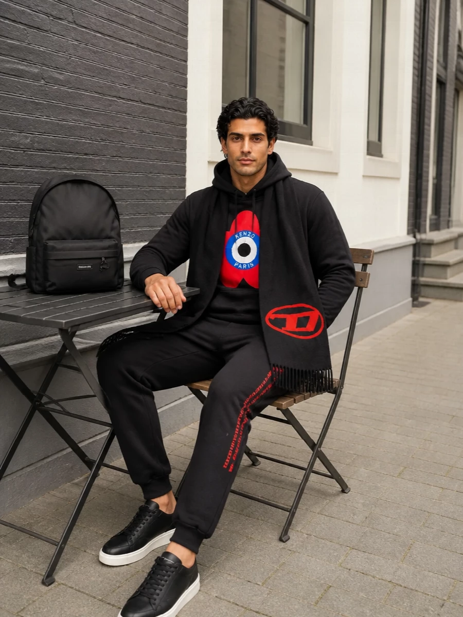 A graphic Kenzo hoodie sits over sharp black joggers for a clean, street-sport base with the red detailing quietly echoed in the side logo. Black low-top sneakers and a streamlined designer backpack keep everything grounded and practical, so the bold flower emblem reads intentional rather than loud. The logo scarf pulls the red accents together for colder days without breaking the monochrome foundation.