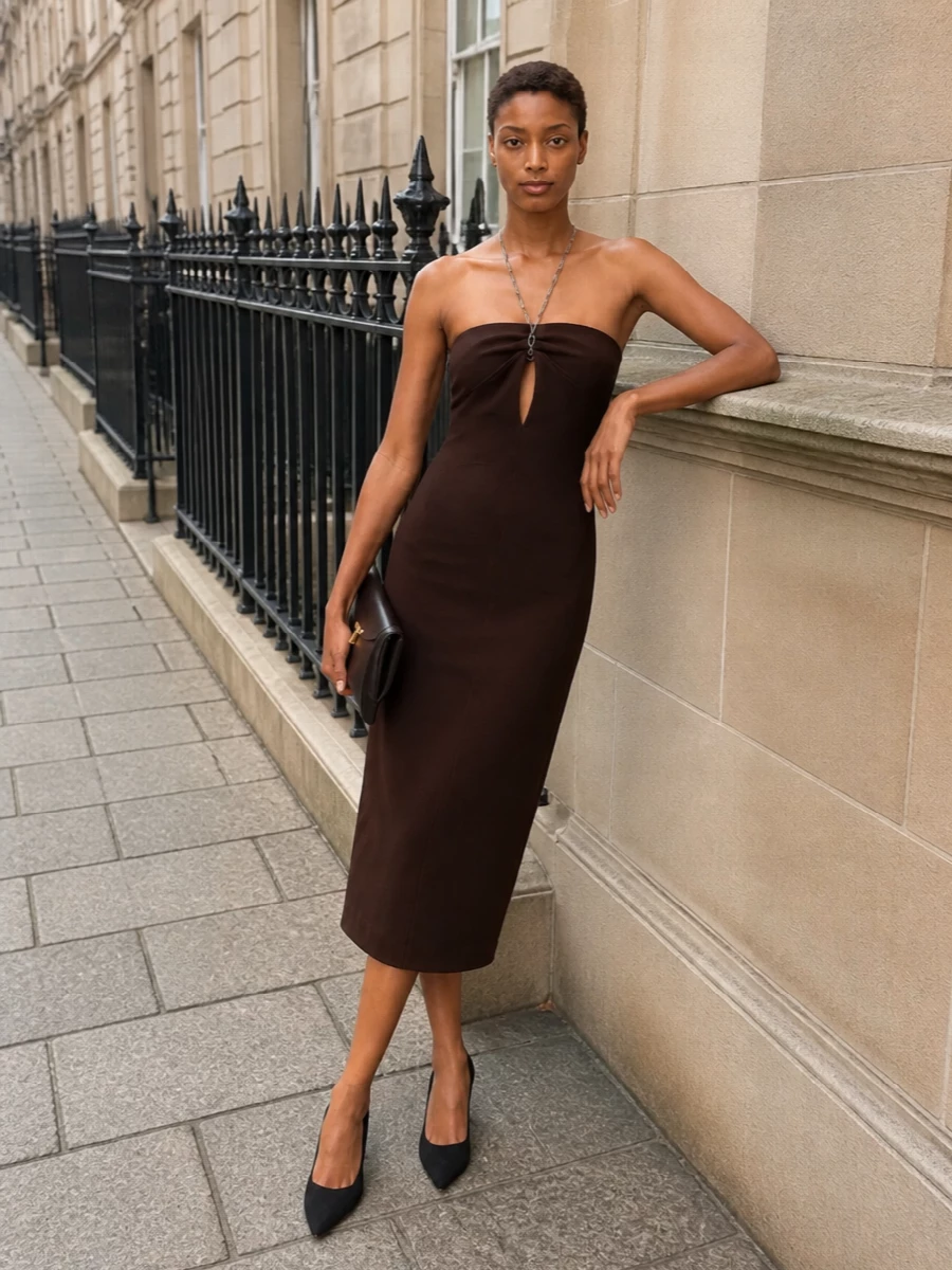 A deep chocolate halter midi and razor-clean black pumps frame the Toteme clutch in a strictly pared-back palette of brown, black and gold. The result is a long, uninterrupted evening line where the built-in chain detail and sculpted bag do all the talking. Choosing the dress route keeps the mood sharper and more minimal than tailoring, ideal for dinners and after-dark rooms that call for quiet confidence.