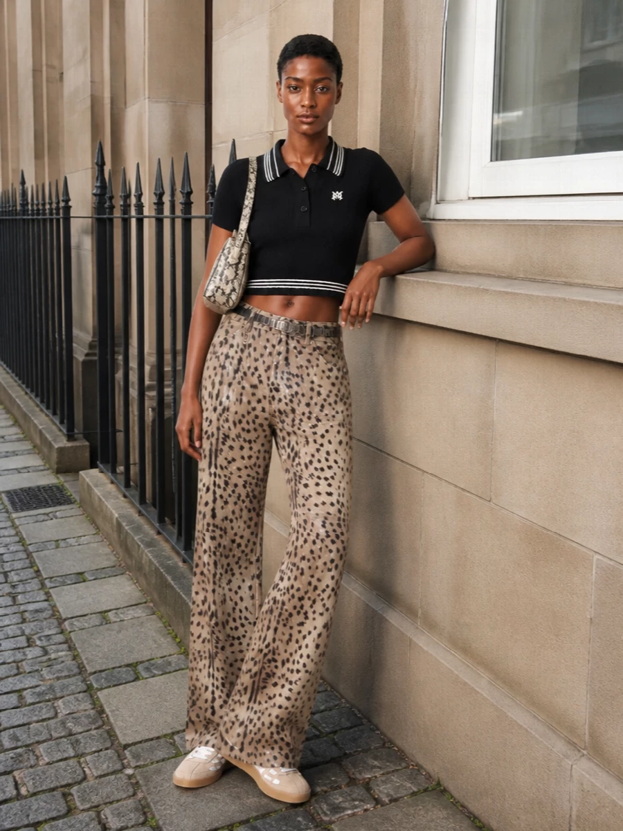Sand toned animal-print denim takes the lead, grounded by a cropped black knit polo and neutral suede sneakers for a clean, athletic street attitude. The wide leg and shorter top play up proportion, while a stone snake-print shoulder bag and slim metal-detailed belt add just enough texture and shine to keep the palette rich but controlled. It feels built for late afternoons that slide into night, with the print doing the talking and the lines kept sharp and unfussy.