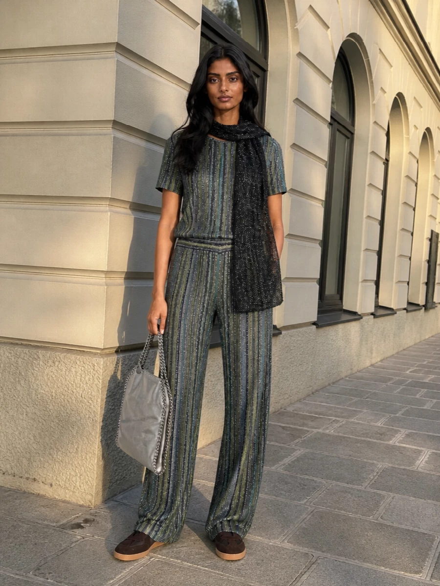 A head-to-toe Missoni knit story keeps the eye moving in vertical lines, with the fluid striped tee and matching wide-leg pant reading like an elongated, modern column. Deep greens, teals and charcoals are anchored by rich brown sneakers, while a silver-grey chain bag and subtle monochrome scarf echo the shimmer for city evenings that skew relaxed yet polished.