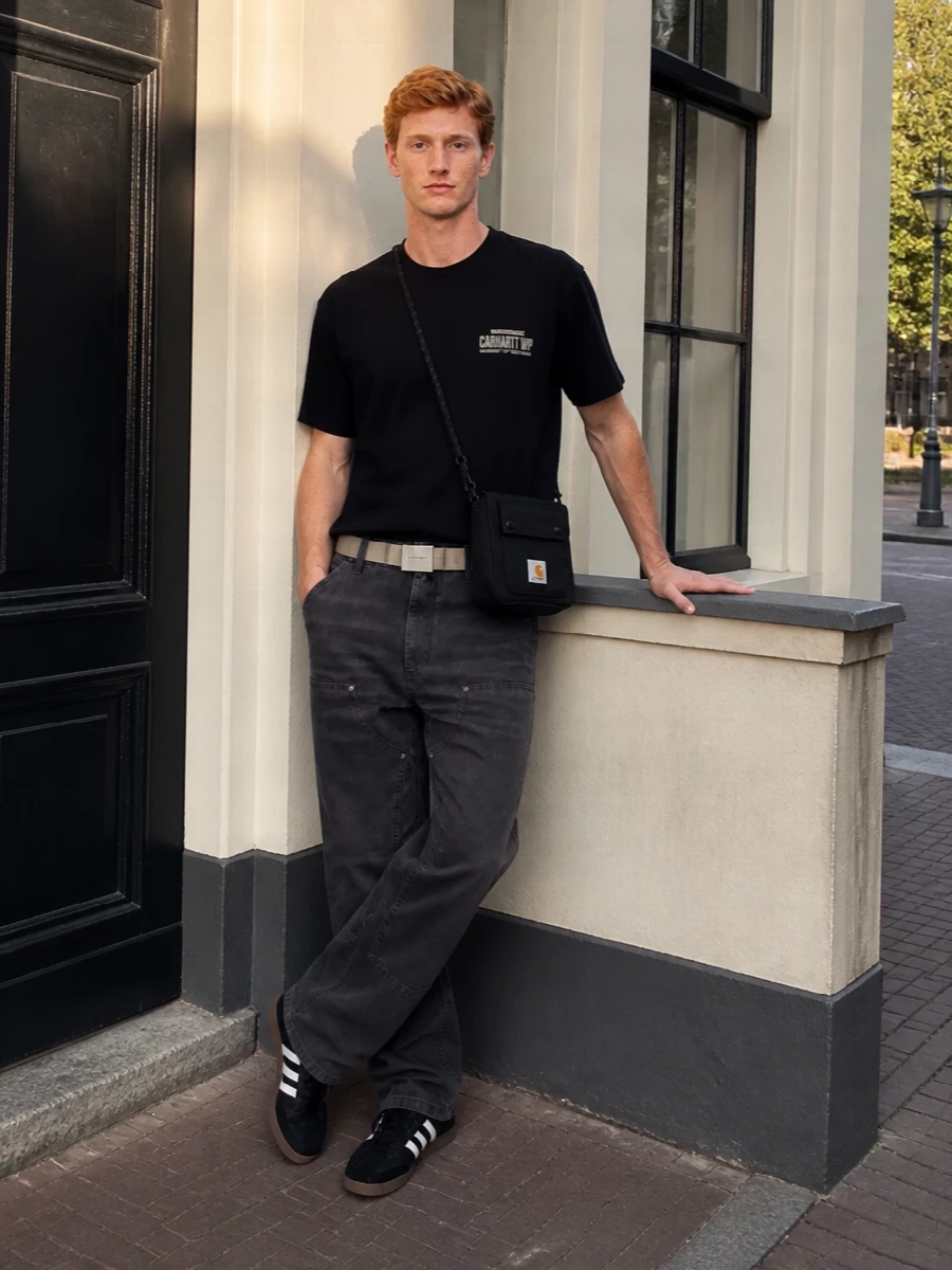 Head-to-toe Carhartt keeps this grounded in clean streetwear, with the black logo tee sitting over washed charcoal work pants for an easy, skate-adjacent base. Classic black-and-gum Adidas sneakers pick up the low-key graphic energy without shouting. A compact black crossbody and sand webbing belt finish it with practical utility and just enough hardware to feel considered, not busy.