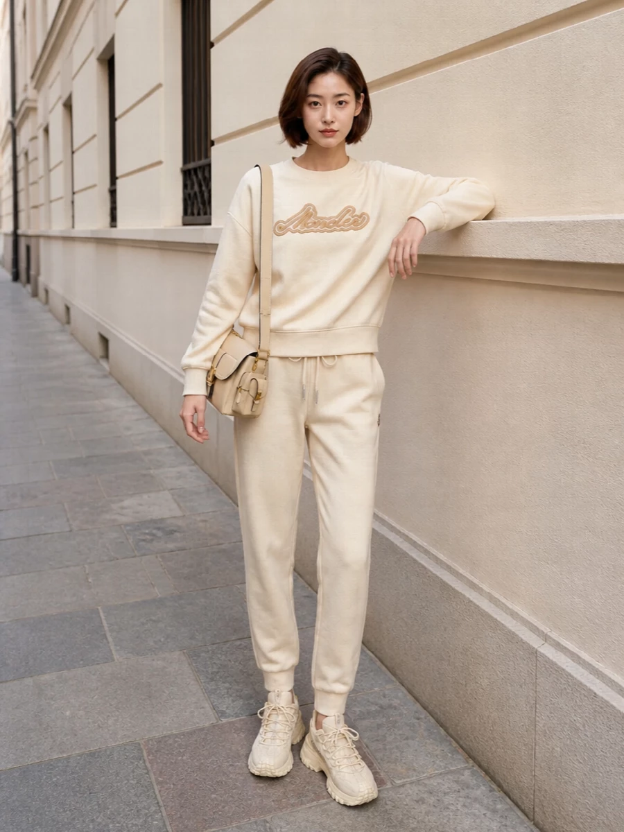 This look leans into a monochrome buttercream story: the relaxed Moncler joggers and matching logo sweatshirt form an elevated tracksuit that feels plush yet understated. Creamy chunky sneakers keep the palette cohesive while the structured Chloé shoulder bag adds just enough refinement to take the outfit from cozy downtime to polished airport or city errands.