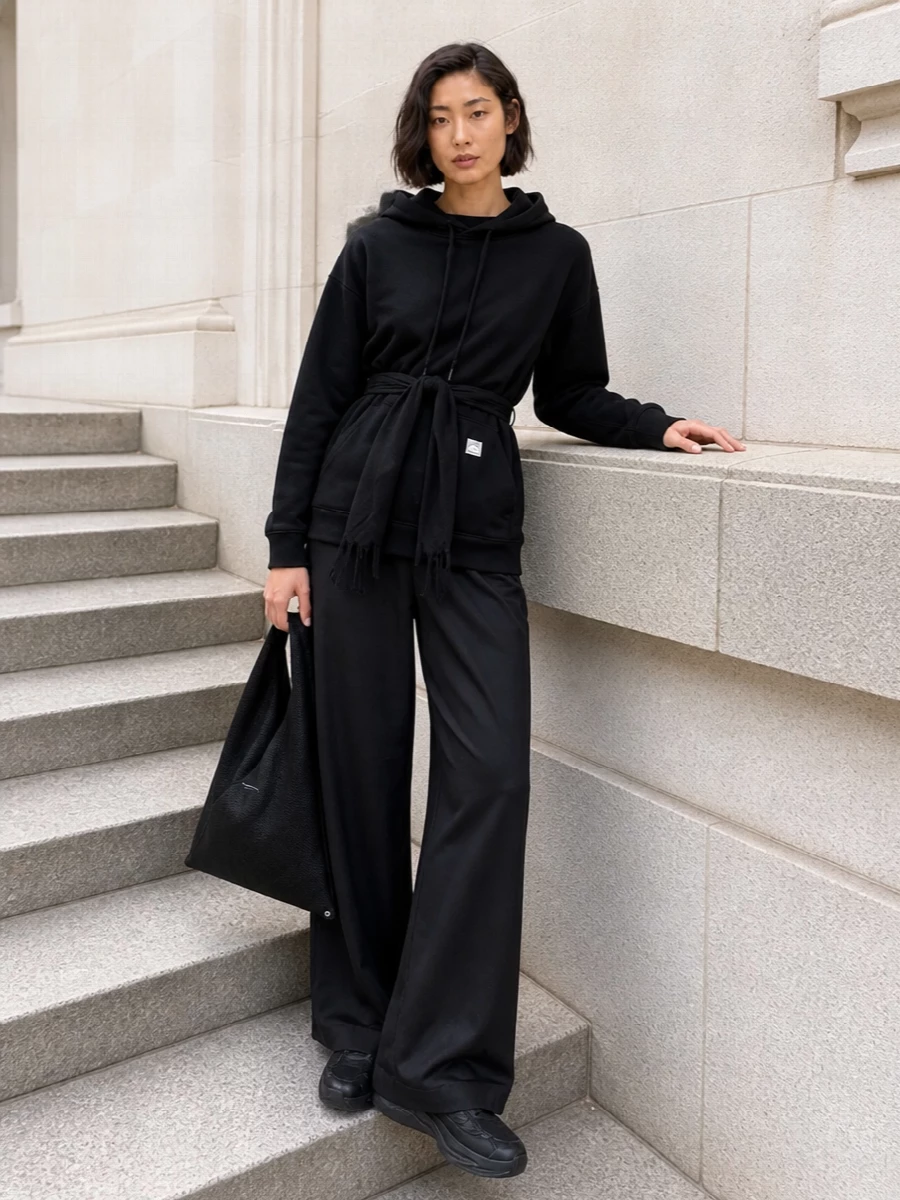 A cinched black hoodie meets inky wide-leg trousers for a monochrome take on streetwear that feels intentional rather than lounge. The all-black runner grounds the volume, while the sculptural mesh tote and quietly branded belt lean into Margiela’s conceptual uniform energy. Head-to-toe, it reads like a city-ready set built from pure shape and texture, not noise.