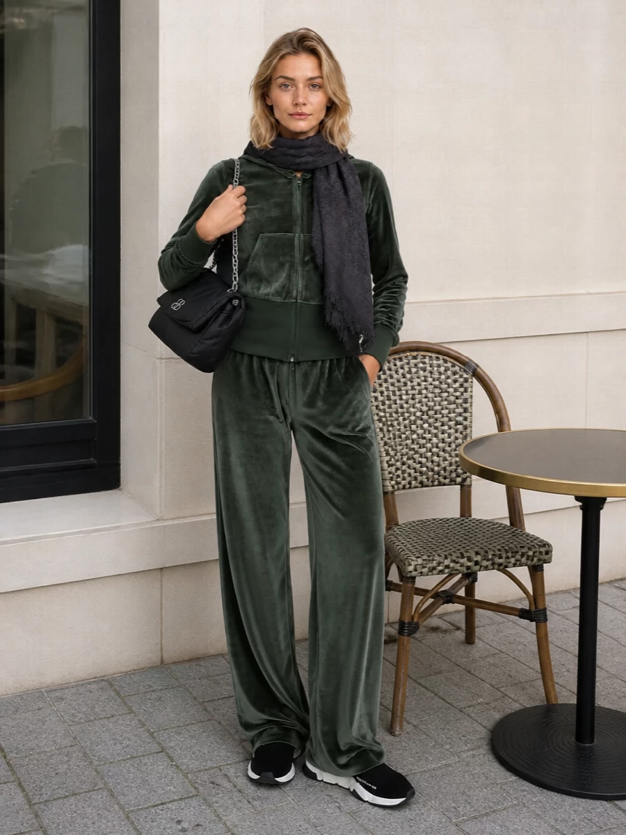 Deep moss velour runs head to toe, so the hoodie and matching wide-leg pant read as a single, fluid statement set. Black sock sneakers and a padded shoulder bag keep it grounded and graphic without breaking the palette. A dark charcoal scarf introduces just enough texture and quiet logo detail for cold city days while maintaining the minimalist, street-sport attitude.