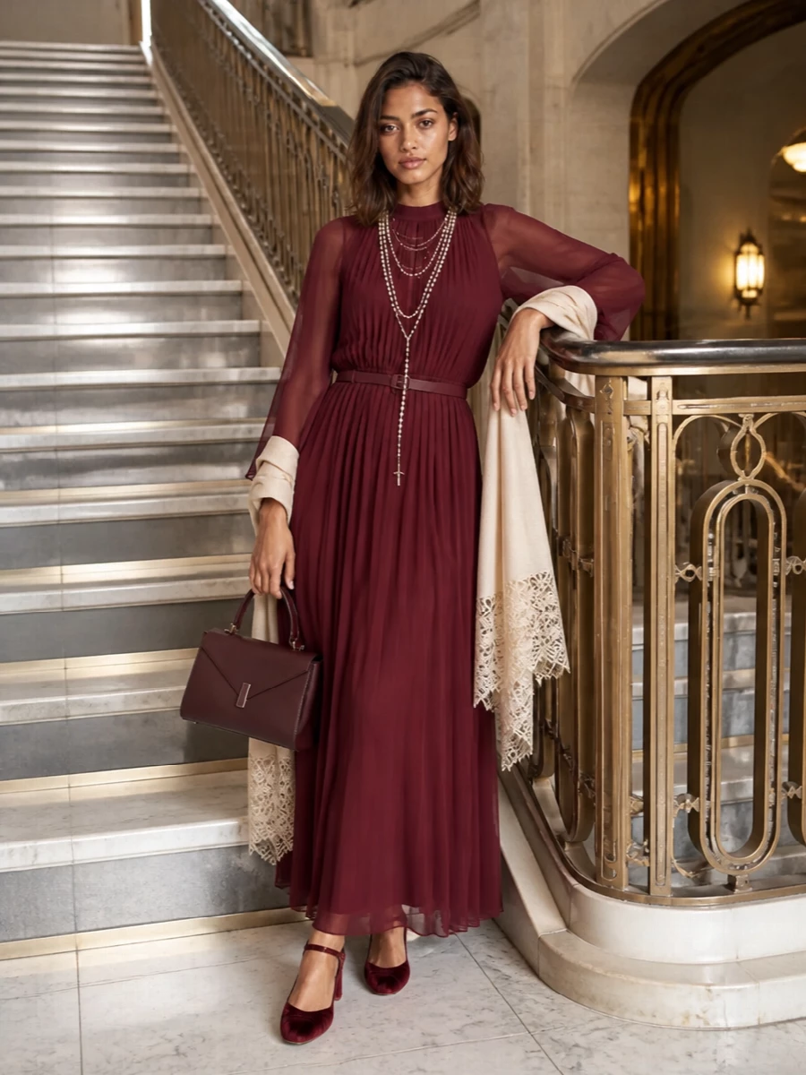 A deep wine column of pleats stays centre stage, framed by tonal velvet heels and a sculpted burgundy top-handle that keep the mood rich but controlled. Pearl draping and a soft lace wrap bring just enough light and texture to make the high neckline and sheer cape sleeves feel intentional for winter evenings. The result is a long, uninterrupted line that reads regal, romantic and quietly assured rather than overtly showy.