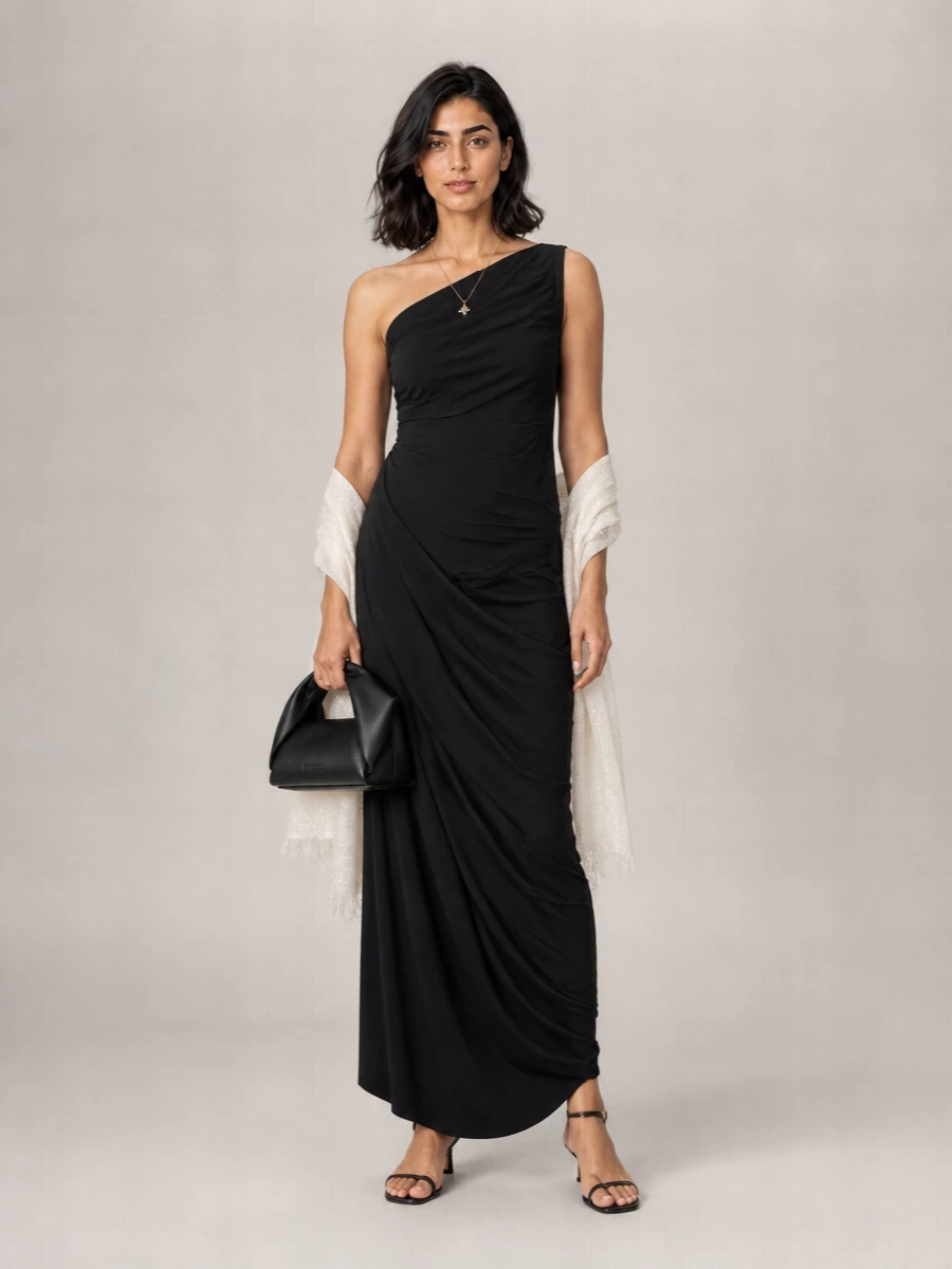 An asymmetric black maxi dress is paired with barely-there black kitten-heel sandals and a sculptural black top-handle bag for a clean, directional evening ensemble. A single gold pendant and an ivory shimmer wrap add just enough light and softness, keeping the mood modern and quietly dramatic.