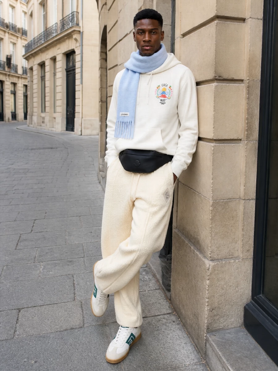 This look leans into Casablanca’s tennis-club nostalgia with an off‑white graphic hoodie paired to tonal cream wide-leg joggers for a plush, monochrome base. Balmain’s off‑white and teal court sneakers and a compact Moncler belt bag keep the silhouette sporty yet refined, while the ice-blue scarf adds a gentle hit of colour and FW texture without disturbing the clean, relaxed lines.