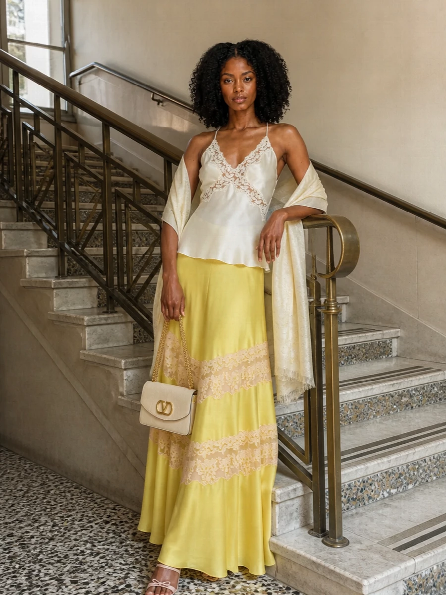 Alberta Ferretti’s butter-yellow lace-panel maxi skirt is paired with an ivory satin camisole for a lingerie-inflected evening ensemble that feels luminous yet restrained. Blush strappy heels and a buttercream Valentino flap bag keep the palette soft and elongated, letting the skirt’s movement stay centre stage. A whisper-light neutral stole finishes the outfit with just enough ceremony-ready refinement for summer weddings or terrace cocktails.
