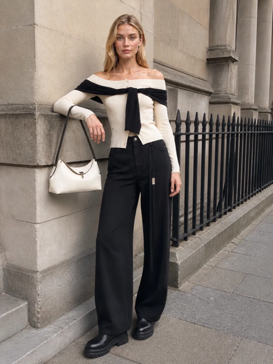 Cream and black stay deliberate here, with the sculptural off-shoulder knit slipping into elongated black wide-leg tailoring for a modern, quietly polished evening mood. Chunky black ankle boots keep it grounded and urban rather than precious, while the cream-and-black shoulder bag picks up the trompe-l’œil tie detail so the palette feels intentional from every angle. It is the kind of outfit that handles a gallery opening, a late dinner and the walk home without a single adjustment.