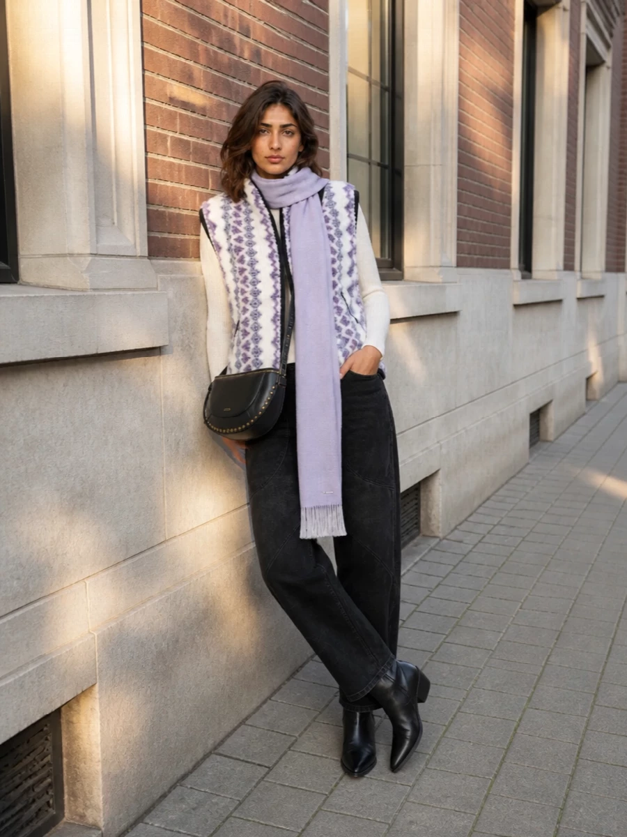 Isabel Marant Étoile’s patterned gilet sits over washed black straight-leg trousers for a grounded, easy take on alpine-inspired boho. Black western boots and a compact black saddle bag sharpen the palette while keeping the mood relaxed. A slim lilac scarf quietly picks up the vest’s lavender and draws the eye vertically, balancing the cozy texture with a clean, elongating line.
