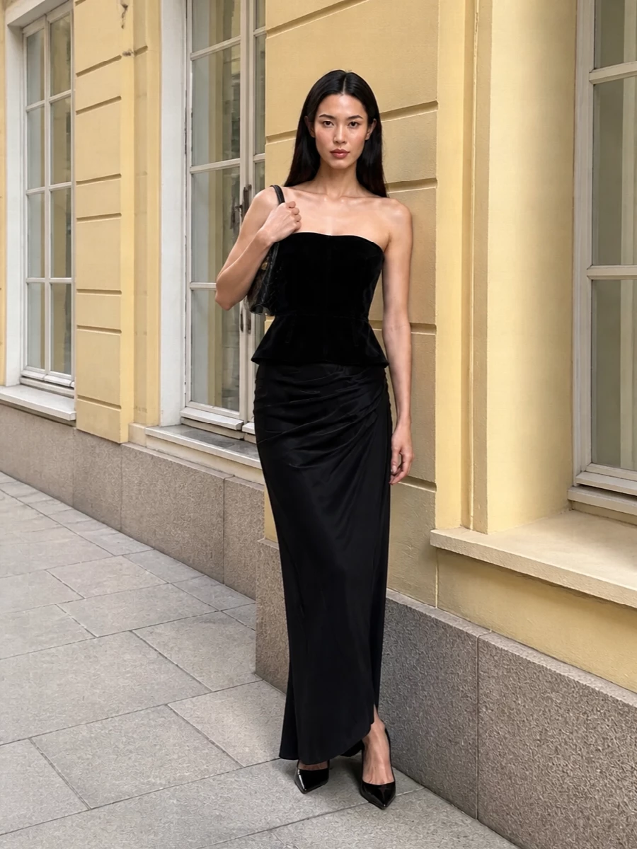 This look is pure nocturnal Tom Ford: a sculpted black velvet strapless top cinching the waist above a liquid, asymmetric floor-length skirt. Glossy patent stiletto pumps and a slim intrecciato clutch keep the line long and graphic, delivering a quietly opulent, all-black evening column that feels razor-sharp yet sensual from every angle.