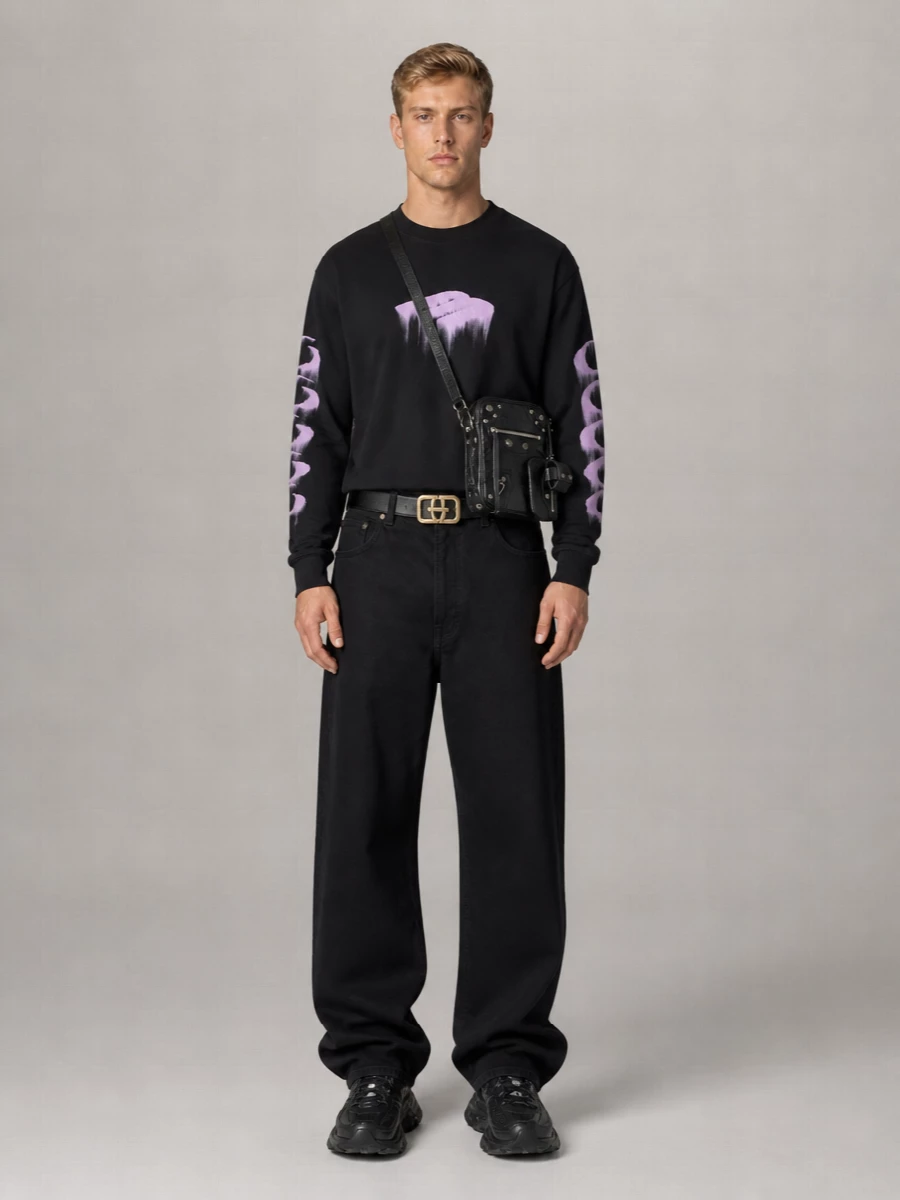A black and lilac graphic tee drops into long, puddling black denim for that signature Balenciaga slouch. All-black technical runners keep the line heavy at the floor, while a compact moto crossbody and sculptural belt buckle sharpen the attitude without breaking the dark palette. The result is gallery-kid streetwear that feels considered but still slightly unruly.