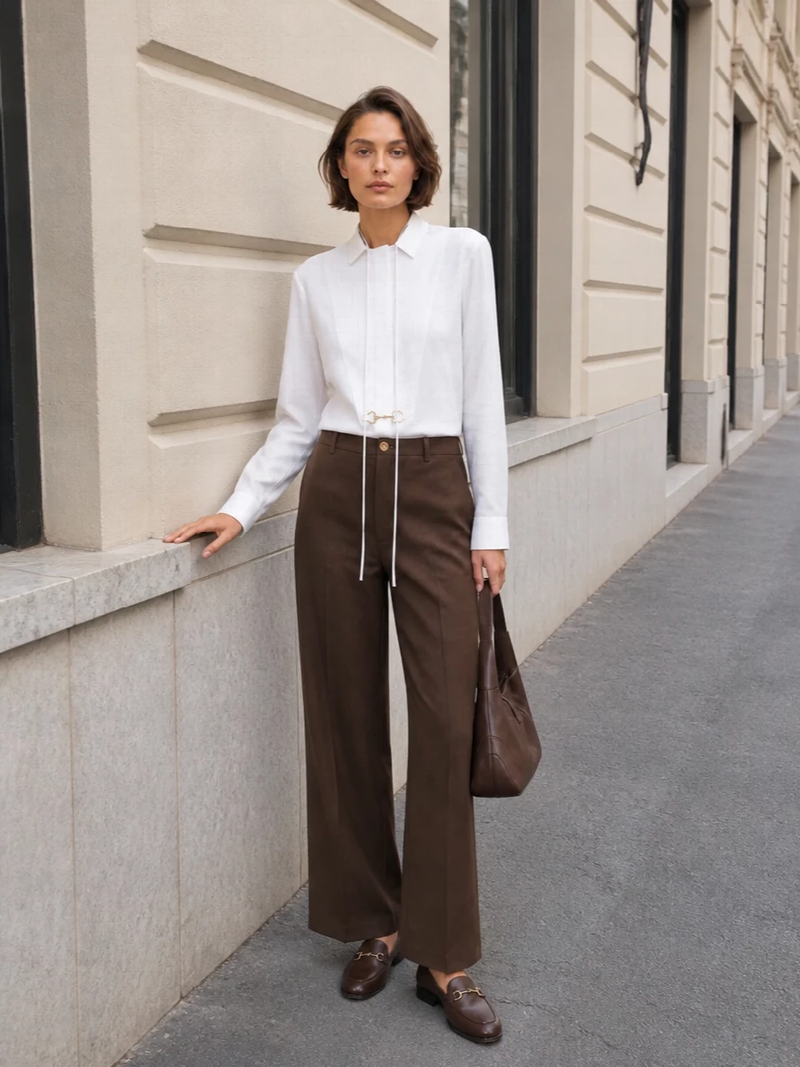 A crisp horsebit-tie white shirt pairs with high-waisted chocolate wide-leg trousers to create a long, disciplined line that flatters the almond-toe loafers. The matching chocolate shoulder bag keeps the focus on subtle texture and tone, delivering a quietly powerful city uniform. This path leans into modern suiting proportions rather than a dress, which better echoes the loafer’s preppy, boardroom-ready attitude.