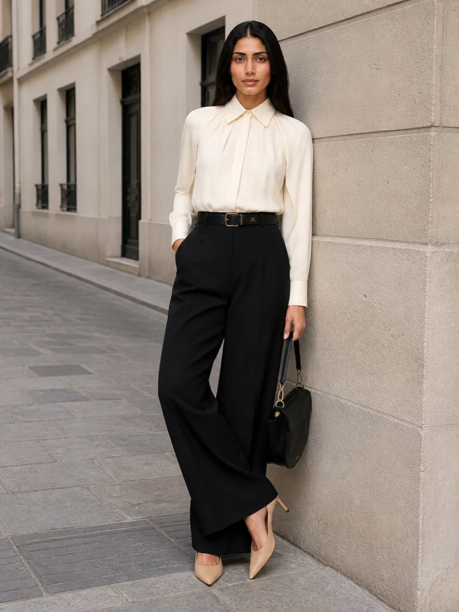 High-waisted black wide-leg trousers are paired with an ivory pleated Max Mara blouse, creating a refined play of structure and softness in a clean monochrome-neutral palette. Nude Prada stilettos lengthen the line, while a sculptural black Wandler shoulder bag and a slim Saint Laurent belt anchor the look with quietly assured, boardroom-to-dinner polish.