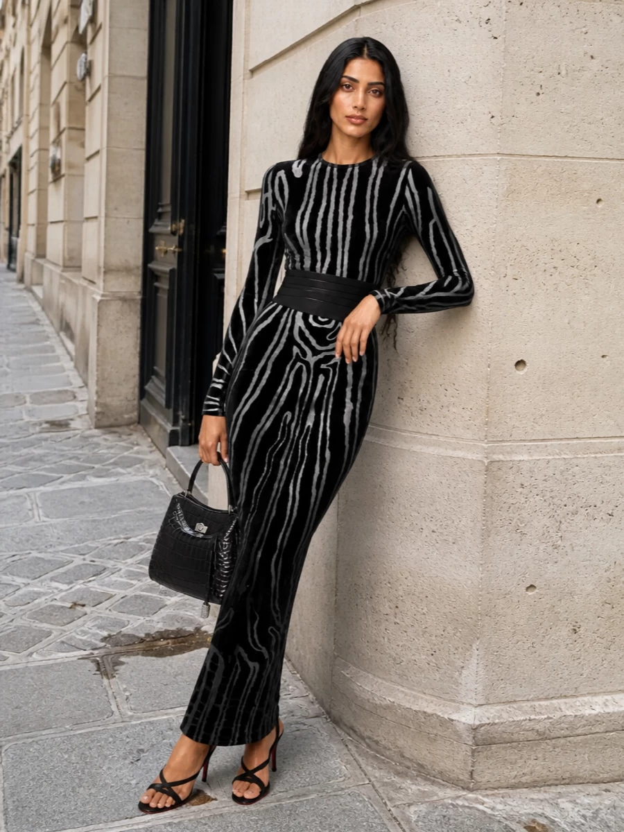This look leans into the dress’s sculpted, woodgrain velvet with a cinched, cummerbund-defined waist and barely-there black strappy sandals that keep the column line uninterrupted. A structured black croc-embossed top-handle bag with silver hardware echoes the dress’s silver tones, delivering a quietly opulent, sharply edited evening statement that feels modern and impeccably polished for FW nights out.