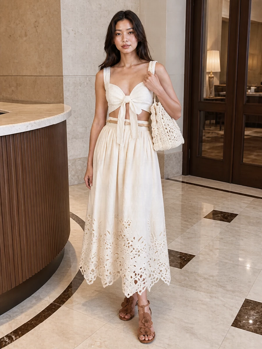 A buttercream cutwork skirt paired with a cropped white tie-front top creates a soft, sculpted waist and a light, romantic line. Tan strappy heels lengthen the leg while a crochet ivory tote and slim braided belt introduce just enough texture to keep the palette interesting without breaking the quiet mood.