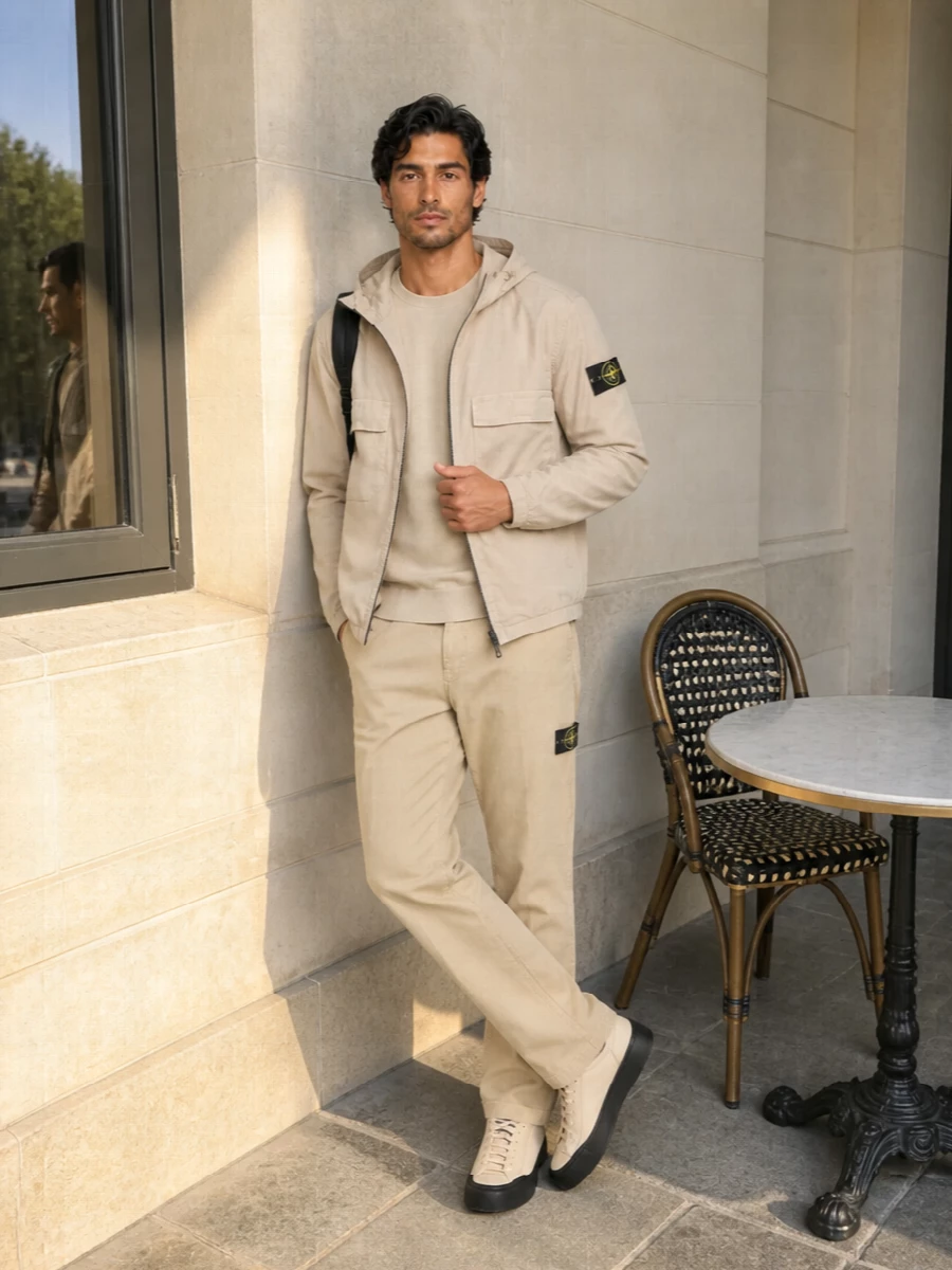 A fully Stone Island neutral utility story, this pairing keeps everything in the sand and khaki family, then sharpens it with black accents from patches, backpack and sneaker sole. The straight-leg chino and regular crewneck sit clean under the technical hooded shell, so the proportions read streamlined rather than bulky. It is city-ready spring dressing that feels quiet, functional and cohesive from head to toe.