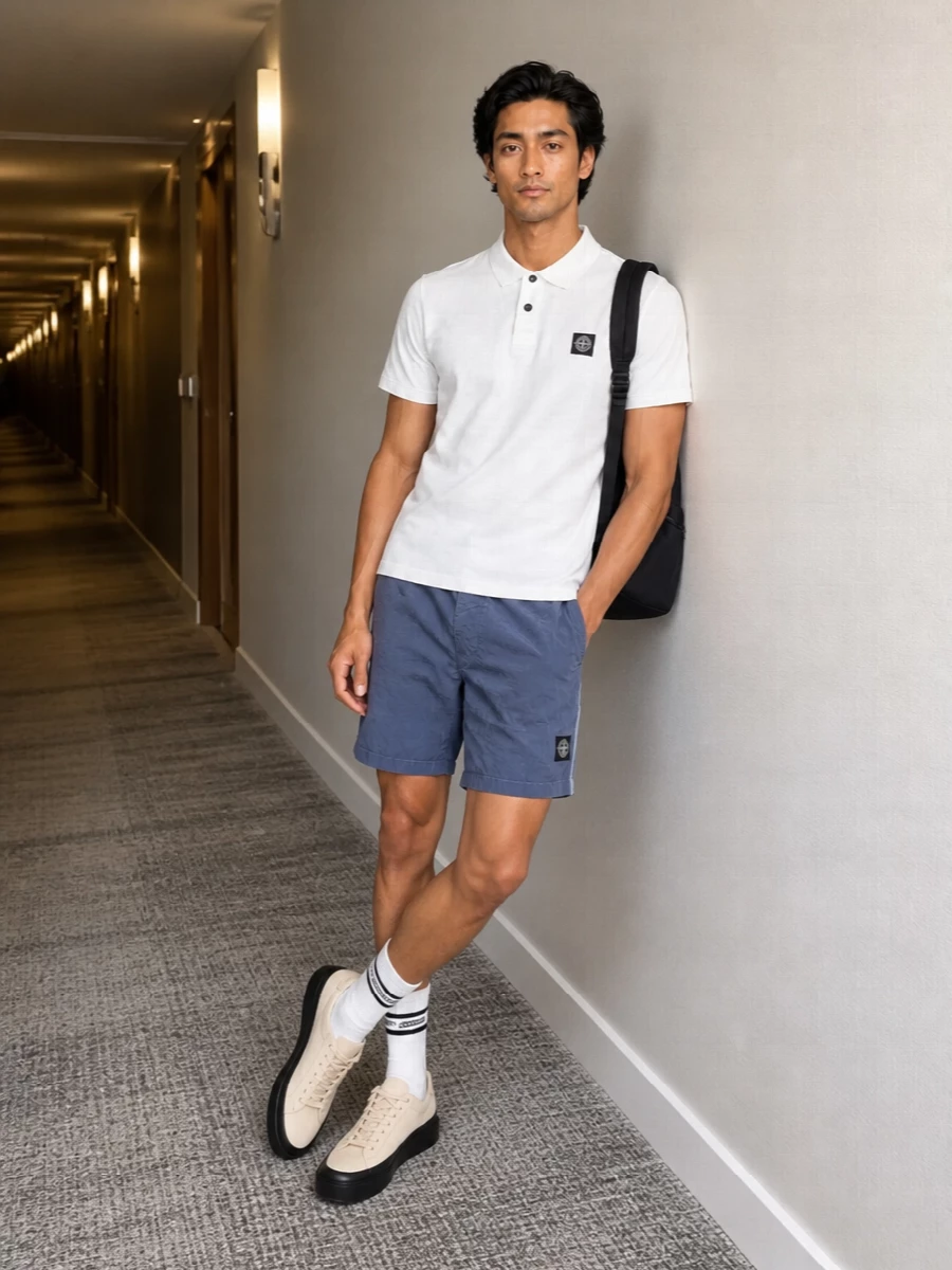 Optic white Stone Island on top, slate technical shorts and neutral sneakers keep this sharply in the sport-street lane without feeling heavy. The palette stays cool and minimal, with soft neutrals and navy grounding the graphic badge details. It is an easy warm-weather uniform that reads clean, athletic and quietly branded from head to toe.