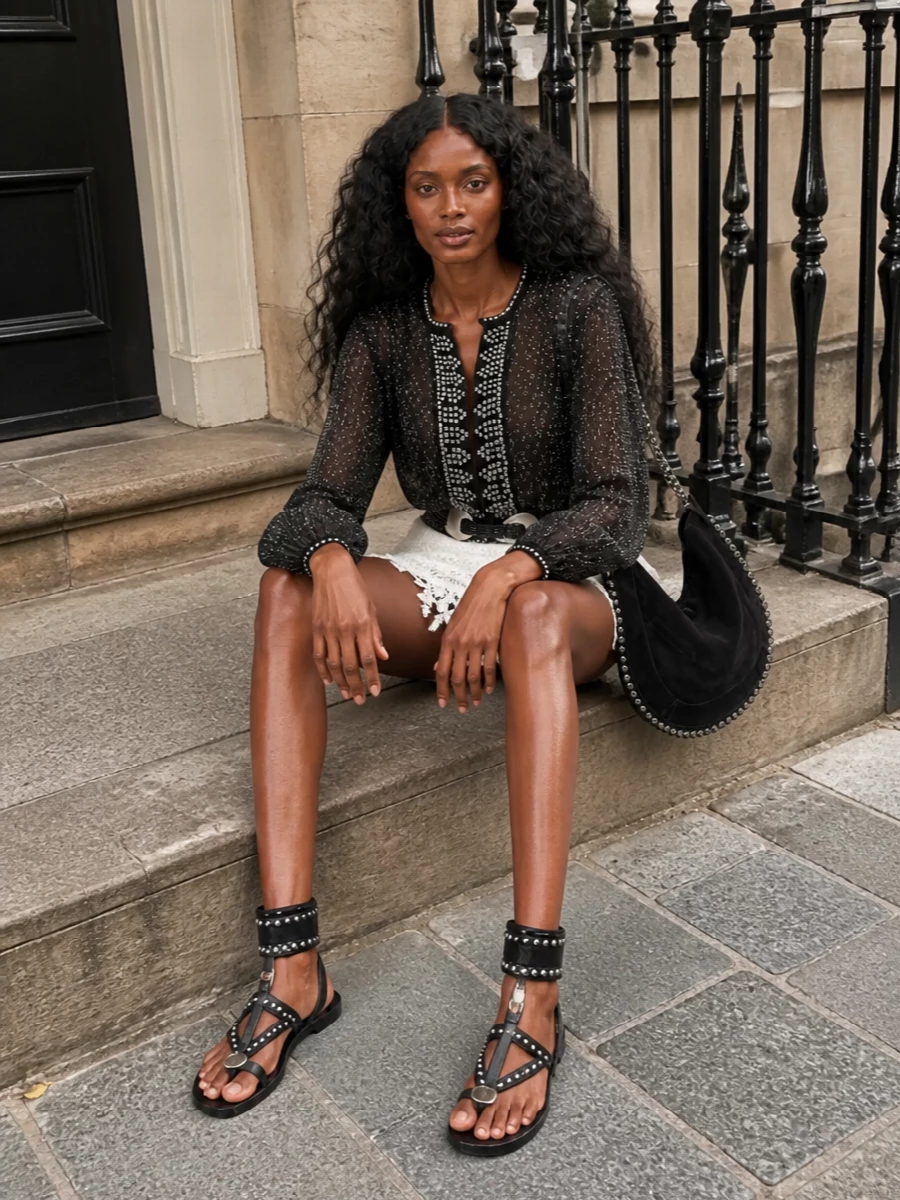 Sheer black embroidery and silver studs skim over a scalloped ivory lace mini for a boudoir-meets-boho evening mood. Flat gladiator sandals and a studded crescent hobo keep it grounded yet glinting, while a slim wrap belt reins in the volume just enough. The result is light, leggy and insouciantly dressed-up for warm nights out.