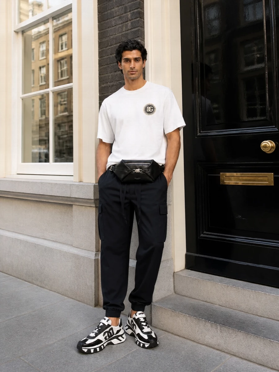 Monochrome and graphic, this outfit leans into Dolce&Gabbana’s logo sport energy while staying clean and considered. The relaxed white tee and technical black cargo joggers frame the chunky sneakers, keeping the focus on proportion and texture. A low profile leather belt bag finishes it with hands‑free ease and a sharp city attitude.