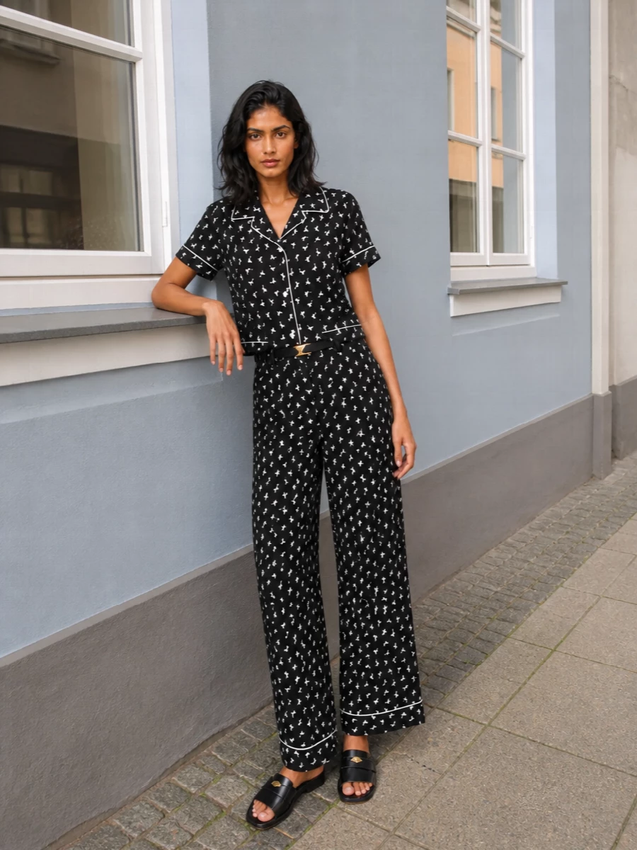 Head-to-toe micro print turns this into a Moschino pajama suit with attitude, the wide-leg trouser and boxy shirt reading more streetwear than sleepwear. Sleek loafer-style slides and a structured logo tote ground the monochrome pattern, while a slim gold-detailed belt adds a precise touch of polish. Wear it for days when you want comfort to feel sharply intentional and a little irreverent.