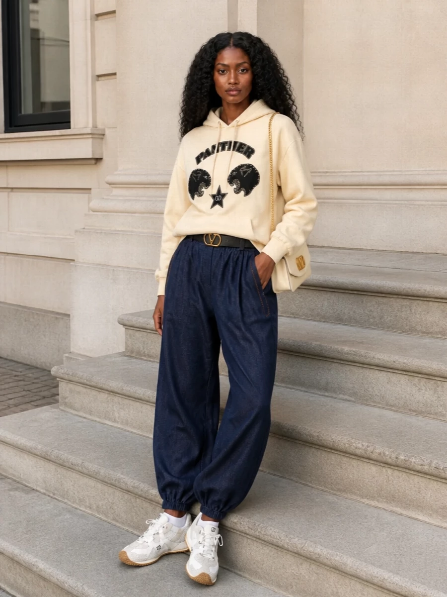 A buttercream graphic hoodie meets dark indigo joggers for a relaxed, sport-led silhouette that still feels sharply edited. Off-white technical sneakers keep the palette soft and grounded. A buttercream shoulder bag and discreet logo belt pull the neutrals together, giving the outfit a polished, streetwise finish.