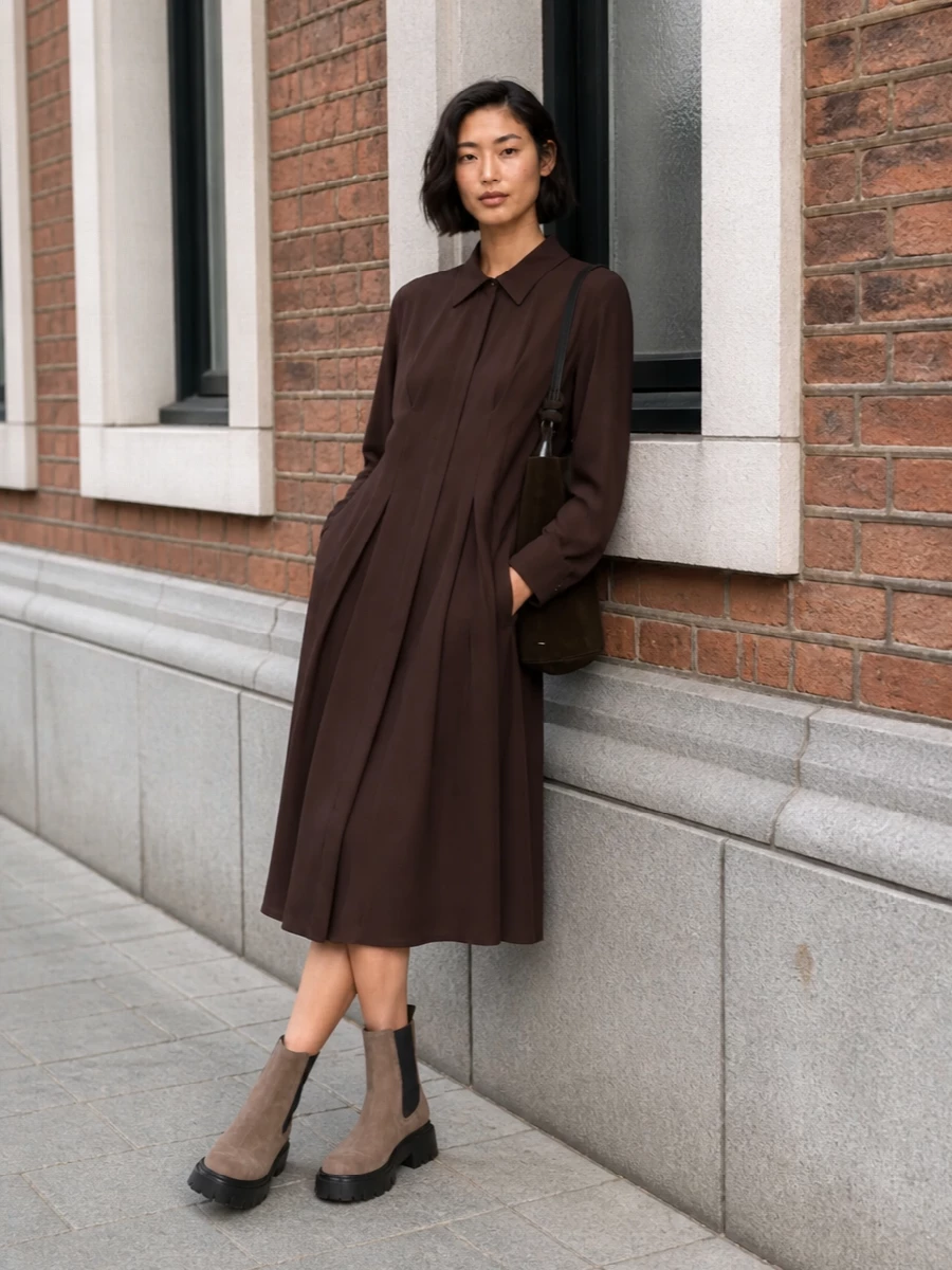 A chocolate pleated shirt dress pairs with the taupe suede Chelsea boots for a grounded, minimalist city uniform. The fluid midi line softens the boots’ chunky tread, while the rich browns stay quietly polished against winter light. A dark suede bucket bag finishes the palette with tactile depth and just enough structure for desk and dinner plans.