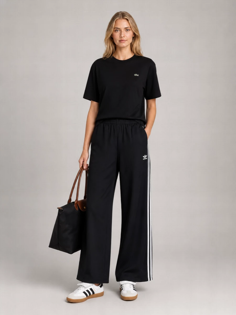 A pure black Lacoste tee meets wide-leg Adidas track pants for a relaxed, monochrome take on sport dressing. White and gum Adidas sneakers and a structured Longchamp tote keep it city ready, while graphic crew socks quietly underline the athletic attitude.