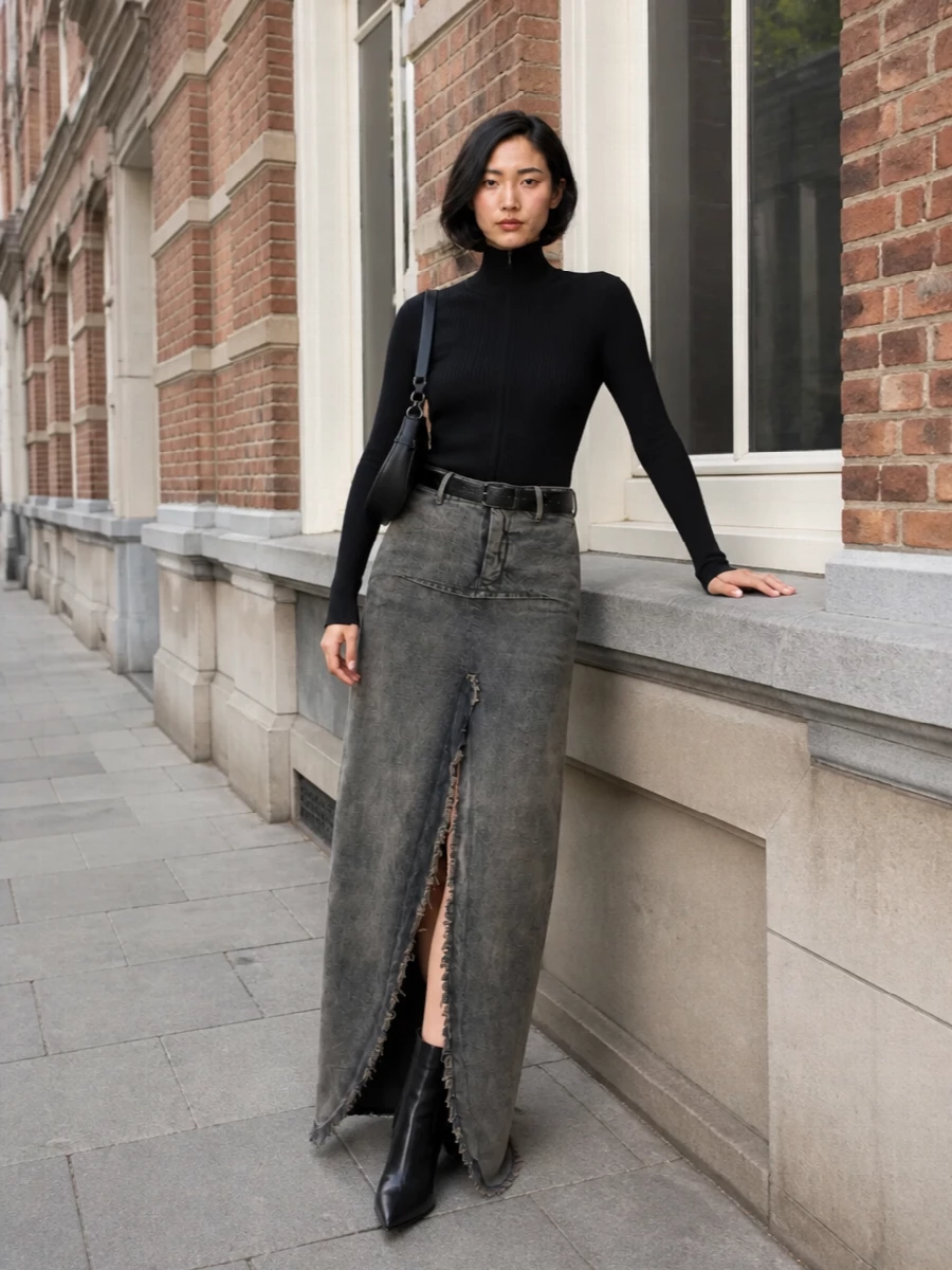 A ribbed high-neck knit seals the torso into a clean black column, letting the frayed denim maxi carry all the drama through that shredded front slit. Sculptural pointed boots and a small crescent shoulder bag sharpen the mood, keeping everything in a strict, urban monochrome. A wide black belt quietly carves out the waist so the long proportions feel intentional rather than heavy.