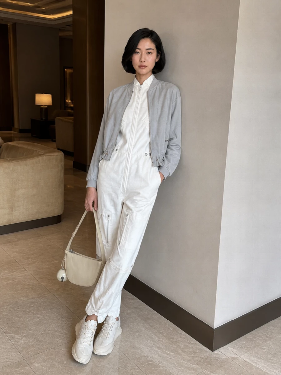 I am anchoring this with the Prada jumpsuit rather than separates, because the one‑piece utility line feels truer to the bomber’s technical minimalism and keeps the silhouette long and lean under its boxy crop. Soft off‑white tones run from shoulders to sneakers and bag, so the suede bomber becomes a gentle contrast in texture rather than color. The result is a quietly confident city uniform with ease of movement and a very considered, modern take on utility dressing.