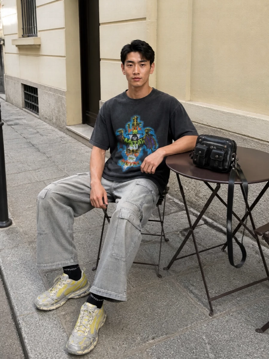 A faded metal graphic tee sits over washed grey wide-leg cargos, keeping the color story muted so the neon artwork takes center stage. Acid-hit runners and a black moto crossbody push the outfit firmly into gritty, late-night streetwear. Logo socks finish it with a small, knowing graphic detail rather than another loud statement piece.