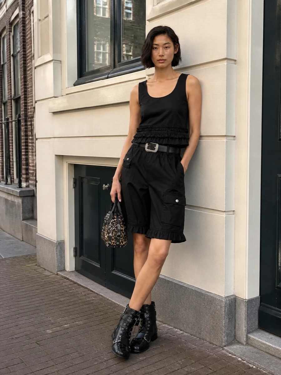 Head-to-toe Ganni in inky black turns the cargo short into a playful, almost co-ord set with the ruffled crop, equal parts sweet and subversive. Lace-up boots and a studded leopard mini bag bring downtown attitude, while the Western-detailed belt quietly cinches everything into a sharp, leggy proportion. It is an easy city uniform with enough texture and hardware to feel styled, not basic.