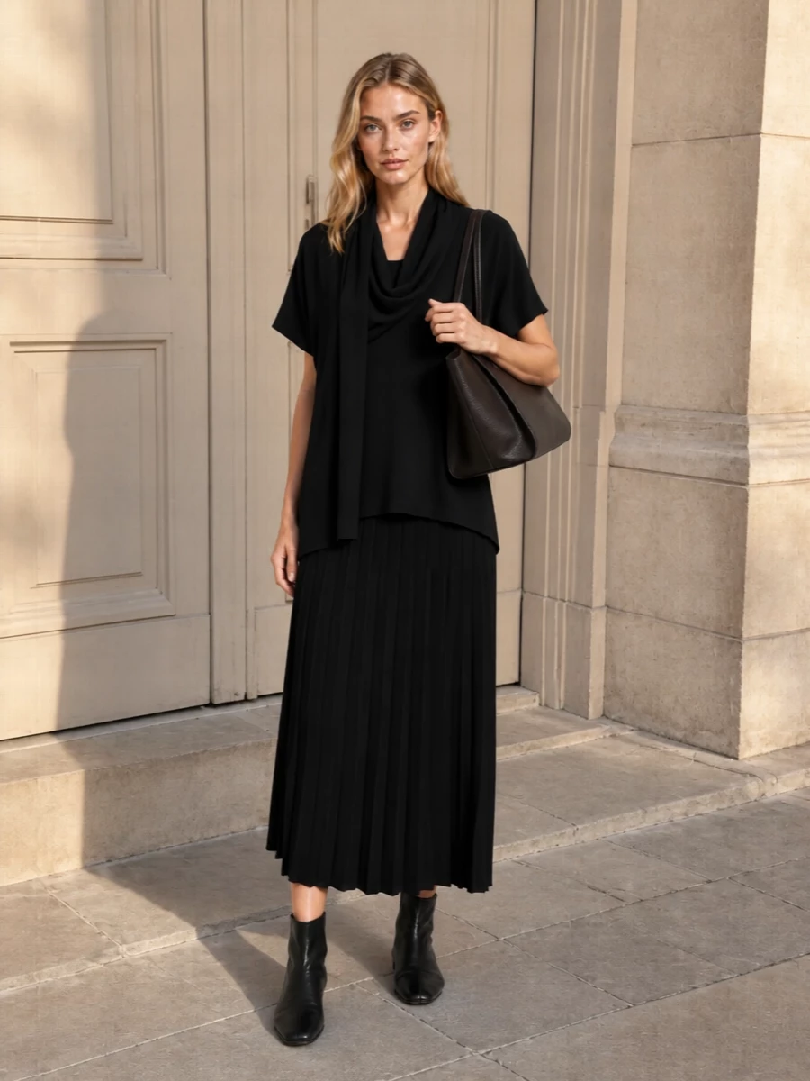 A black knit column that runs from cowl-neck tunic into pleated midi skirt creates a fluid, uninterrupted line that feels calm, modern and quietly confident. Sleek sock boots keep the base sharp but unobtrusive, while the deep espresso tote and clean black scarf add depth without breaking the minimalism. It is an easy uniform for days when you want softness, movement and polish in equal measure.