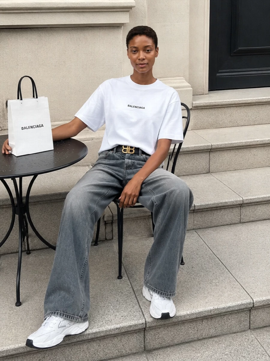 Balenciaga’s white logo tee sits against washed grey wide-leg denim for a clean, slouched silhouette that feels graphic but not shouty. Chunky white runners keep the line grounded and technical, while a sharp white mini tote and slim BB belt bring just enough structure and polish to read as quiet luxury rather than pure athleisure. The result is a monochrome streetwear uniform that looks deliberate yet relaxed from brunch to late city walks.