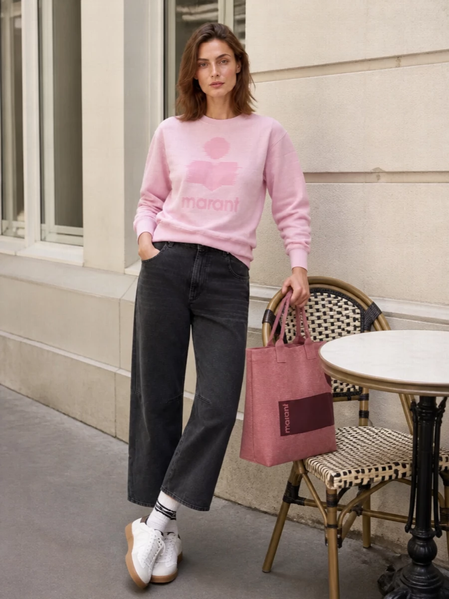 Blush logo fleece meets washed charcoal denim for a soft yet grounded take on Parisian streetwear. White gum-sole sneakers and a dusty rose tote keep the palette light, with logo touches repeating in an easy, off-duty way. The result is relaxed, feminine and balanced, ideal for city days where comfort and polish share equal billing.