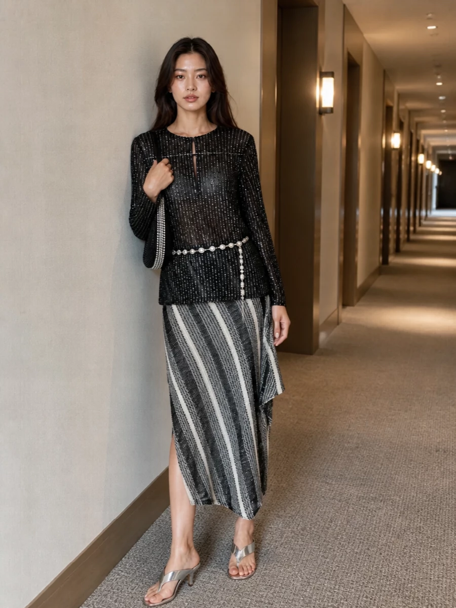 A sheer, sequinned Missoni tunic glows over the monochrome asymmetric knit skirt, creating a long, fluid column that feels sultry yet considered. Mirror-finish silver mules, a crystal-swept shoulder bag and a draped chain belt pick up the sparkle, keeping everything in the same black and silver story without breaking the ease of the silhouette.