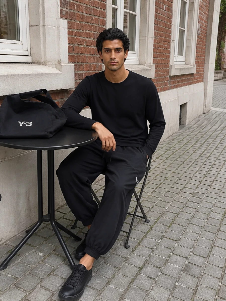 Head-to-toe Y-3 in deep black creates a calm, graphic uniform built around relaxed volume and subtle technical details. The cropped, ballooned track pant plays off the clean long-sleeve, while the all-black low-tops keep the line uninterrupted from shoulder to ground. A soft-structured tote finishes the set with just enough logo to read directional without shouting, ideal for gym-to-city days or travel.