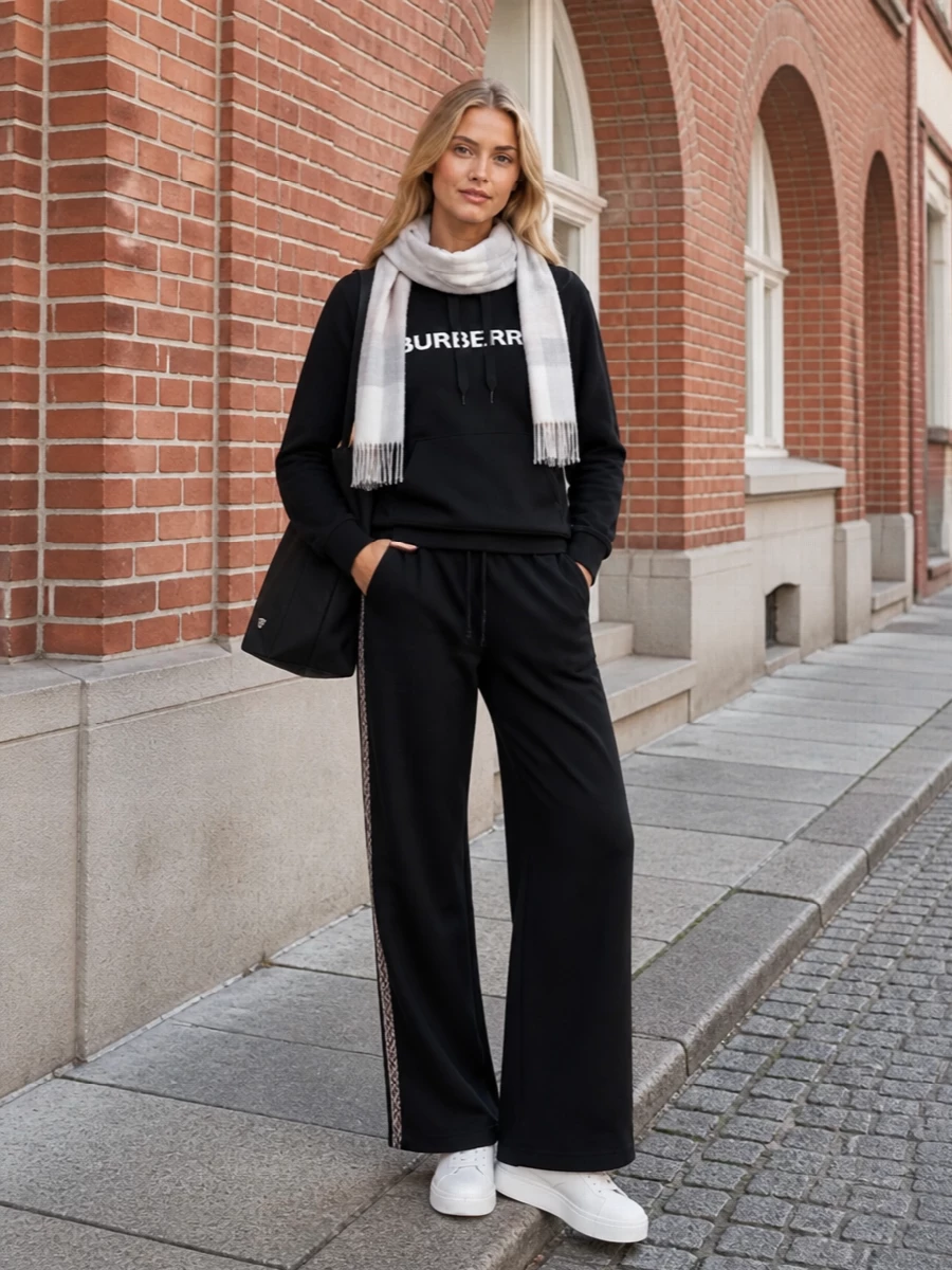 A quietly branded black hoodie sits cleanly over the wide-leg trouser, keeping the mood relaxed but considered. Crisp white sneakers lighten the base and sharpen the sport influence, while the structured black tote leans into a refined travel-ready attitude. The soft neutral scarf adds just enough heritage and warmth without competing with the minimal lines.