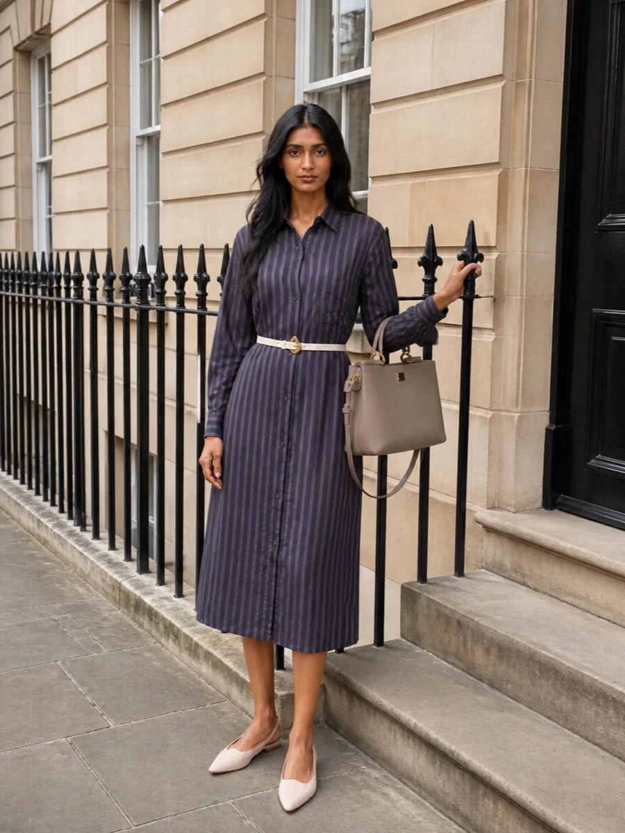 A tailored striped shirt dress cinched with a slim white belt keeps the line long and clean while quietly defining the waist. Blush slingback flats and a structured taupe top-handle bag bring soft, polished contrast that works just as well for a gallery afternoon as for an understated dinner. The mix of inky blue, warm neutrals and a touch of gold hardware reads considered yet relaxed.