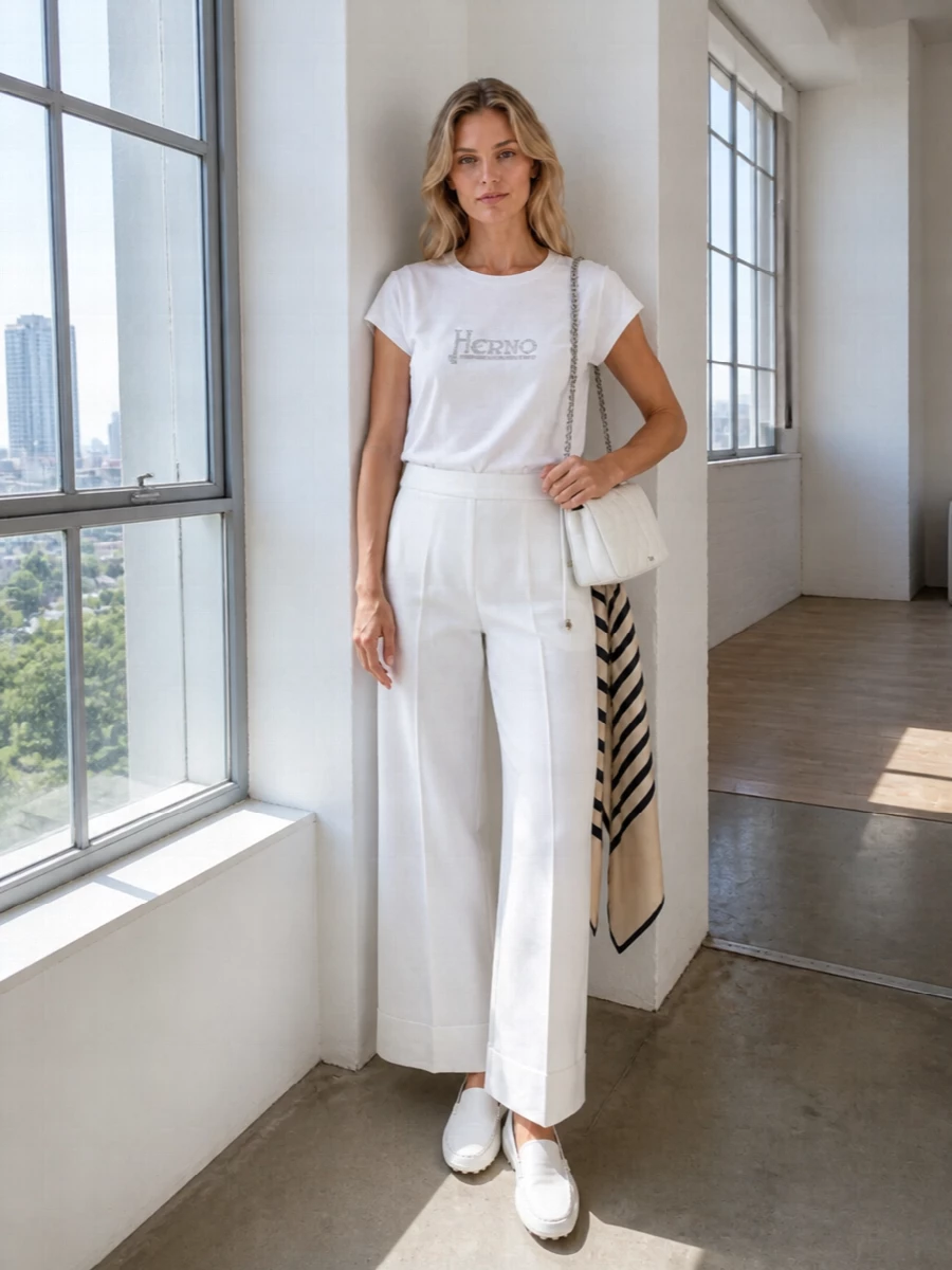 Crisp white tailoring meets relaxed ease, with the crystal‑logo tee tucked into wide-leg trousers for a clean, modern silhouette. Soft-white loafers and a compact quilted shoulder bag keep the mood polished yet quietly relaxed. The graphic scarf adds just enough black to ground the head-to-toe light tones without disturbing the minimalist calm.