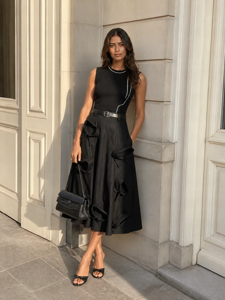 A sculptural black Moschino midi anchors this outfit with couture-level bows, while the sleek crystal-trim shell keeps the monochrome palette intentionally modern for evening. A low black mule and structured black top-handle bag echo the skirt’s polish without fighting its volume, and a slim belt crisps the waist so the A-line silhouette feels sharp rather than sweet.