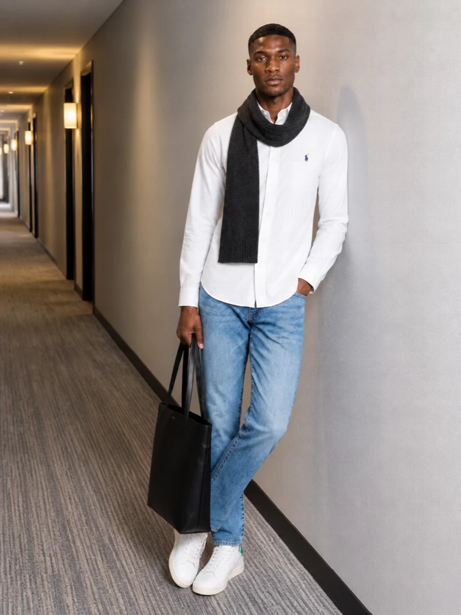A white Oxford shirt pairs with straight-leg light blue denim for a clean, collegiate base that feels relaxed but still put together. Classic off-white court sneakers keep the energy sporty and unfussy, while a structured black leather tote and charcoal scarf add just enough city polish. The result is an easy preppy uniform that works from weekday errands to casual dinners without trying too hard.