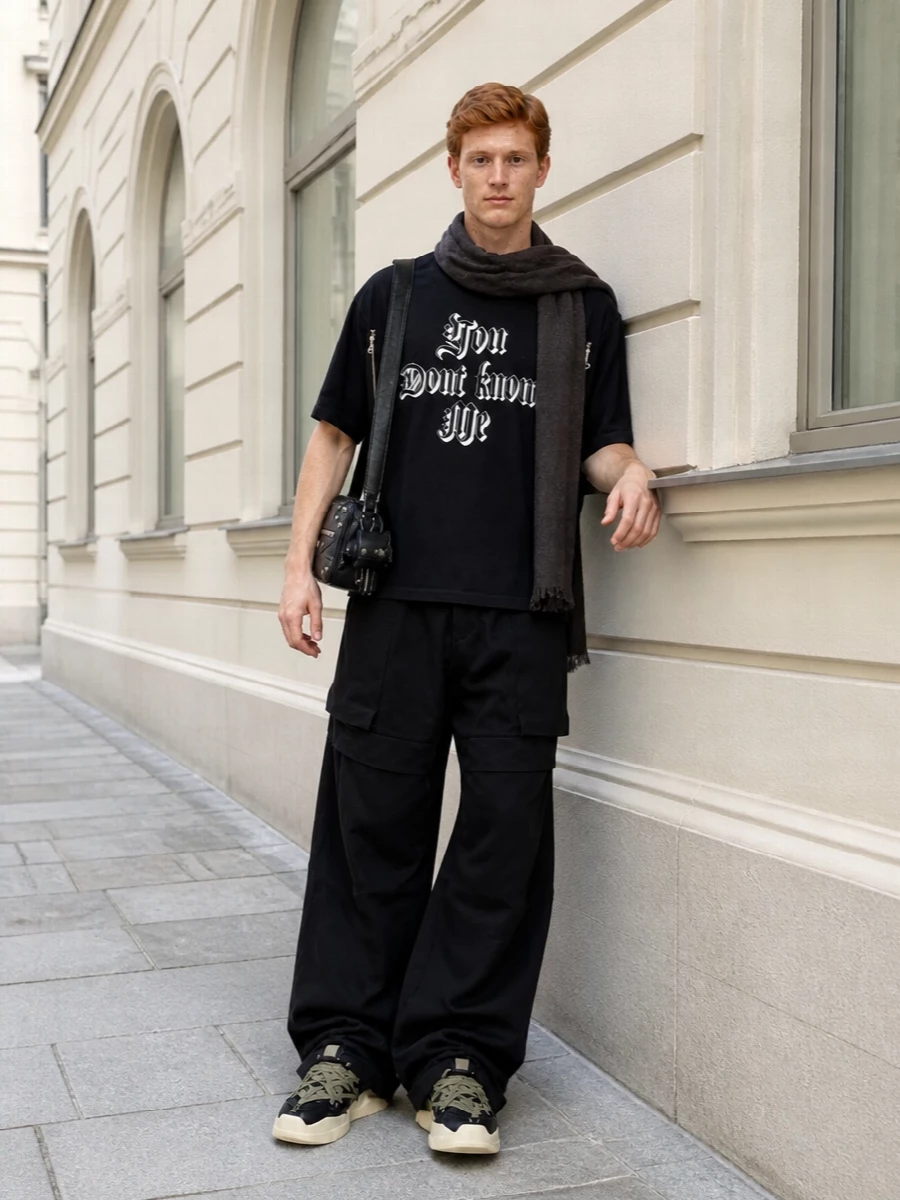 Undercover’s statement tee sits over sweeping Rick Owens cargos for a graphic, shadowy take on utility streetwear. Rick’s high-top sneakers and a hardware-heavy Balenciaga crossbody keep the volume grounded and the mood strictly nocturnal. A dark Rick scarf finishes the frame without diluting the typography or zip detailing on the tee.
