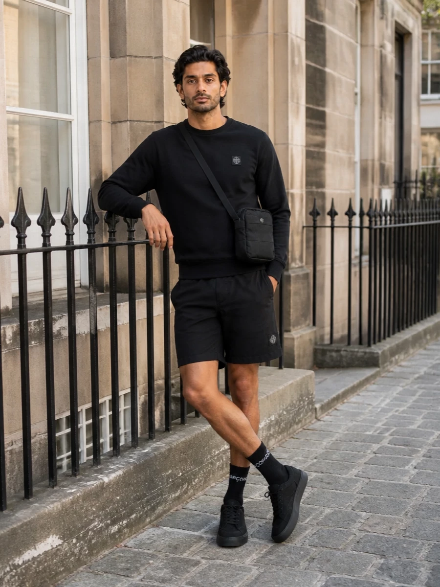 A monochrome Stone Island uniform built around technical black shorts, grounded by a clean black crew-neck sweatshirt and matching low-top sneakers. The compact crossbody and sharp black socks keep everything streamlined and practical for city movement and travel. It reads quietly branded, minimal and sport-led, with just enough structure to feel considered rather than casual-only.
