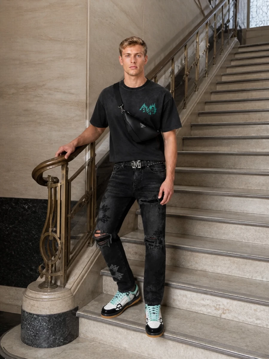 Washed black on black keeps the energy focused on texture and hardware, with the tee’s neon flame hit and the teal sneakers carrying the color story. Straight-leg distressed denim reins in the oversized top so the proportions stay sharp, while the studded belt and clean crossbody add punch and function without feeling heavy. It is a street-led, night-ready combo that reads distinctly Amiri from head to toe without tipping into costume.