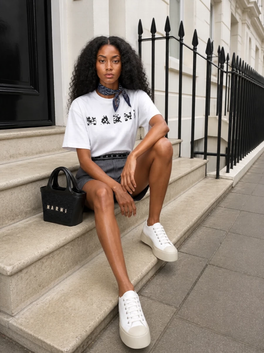 Graphic Marni logos meet sharp suiting-inspired shorts for a fresh, preppy take on streetwear. Chunky white platforms and a black trimmed mini tote keep the palette tight and clean, while a navy silk square adds a subtle collegiate twist without competing with the tee.